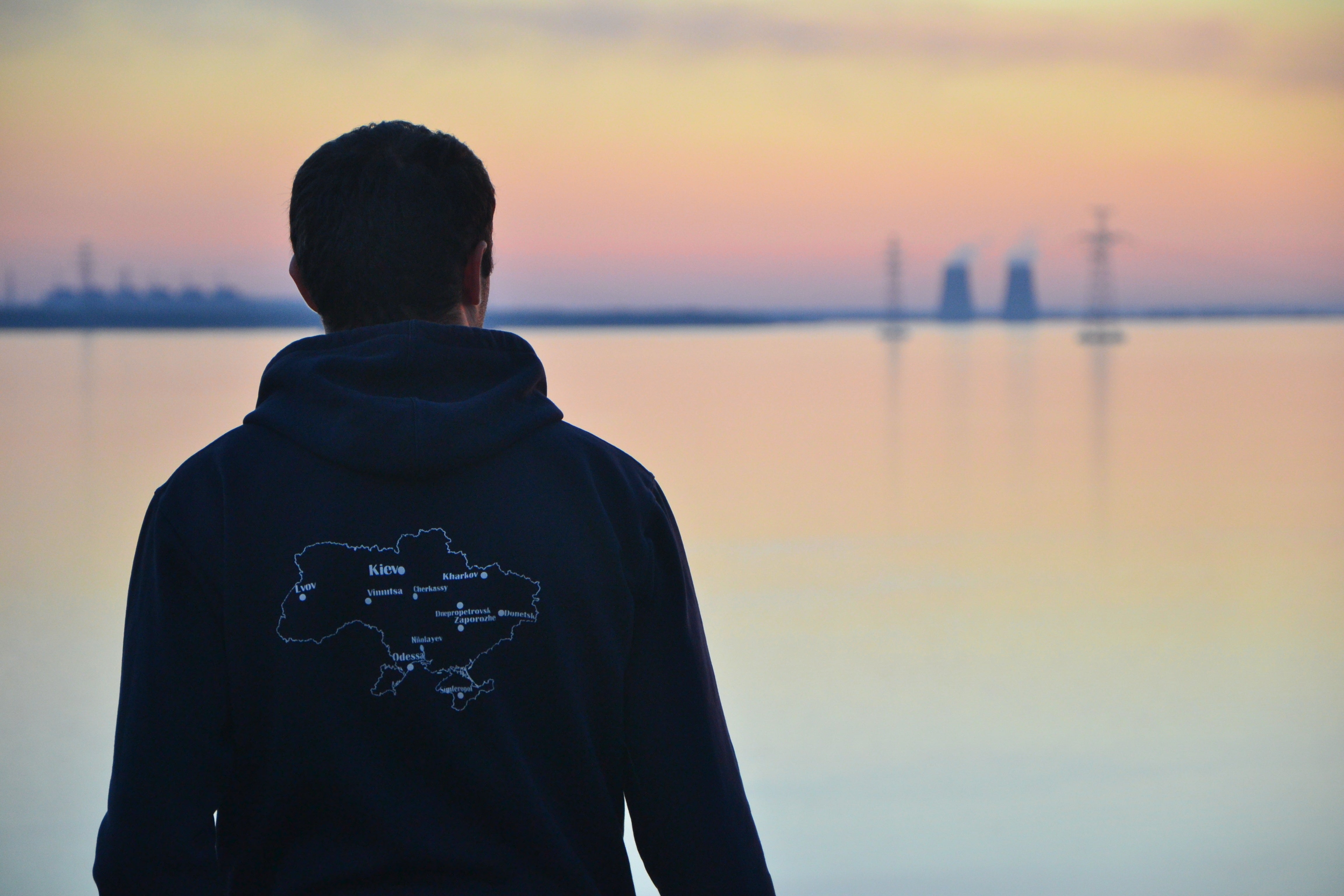 silhouette of a man dressed in a black hoodie with a map of Ukraine standing on the bank of a river and looking at a Zaporizhzhya Nuclear Power Plant at sunset.