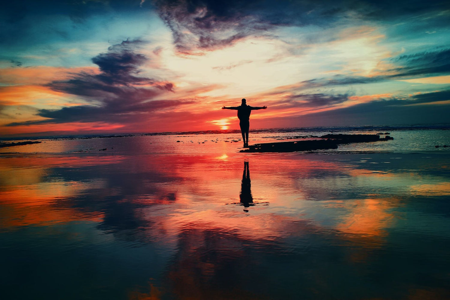 Person standing on the beach during sunset with hands raised in a colorful display.